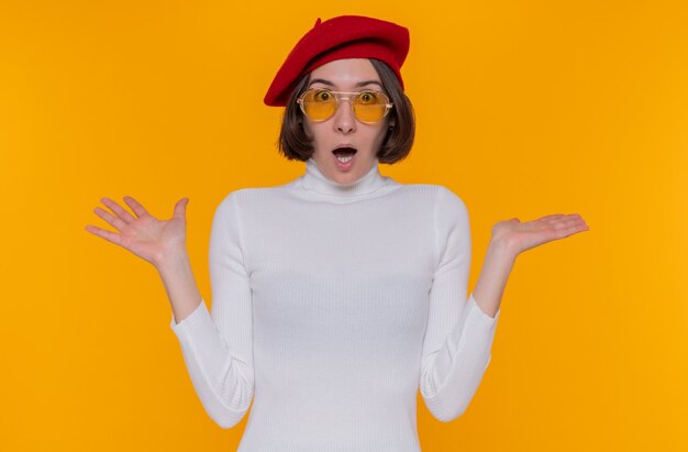 Young woman with short hair in white turtleneck looking at front surprised and confused with arms raised standing over orange wall