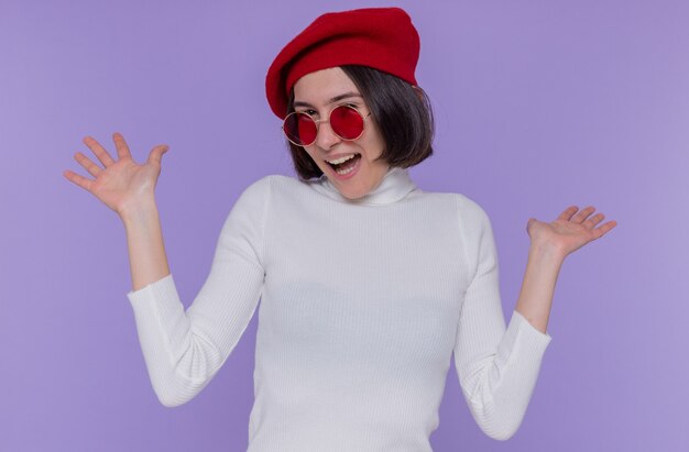 Young woman with short hair in white turtleneck wearing beret and red sunglasses happy and excited screaming rejoicing raising hands standing over blue wall