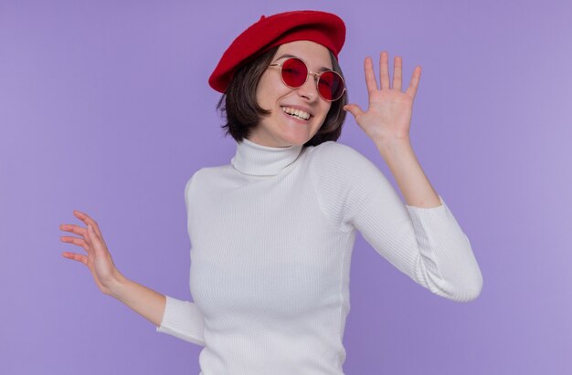 Young woman with short hair in white turtleneck wearing beret and red sunglasses happy and excited smiling cheerfully gesturing with hands standing over blue wall