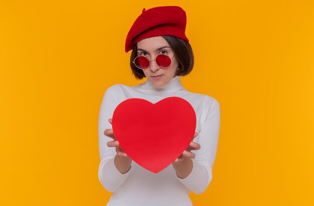 Young woman with short hair in white turtleneck wearing beret and red sunglasses holding heart made of cardboard looking at front smiling confindet standing over orange wall