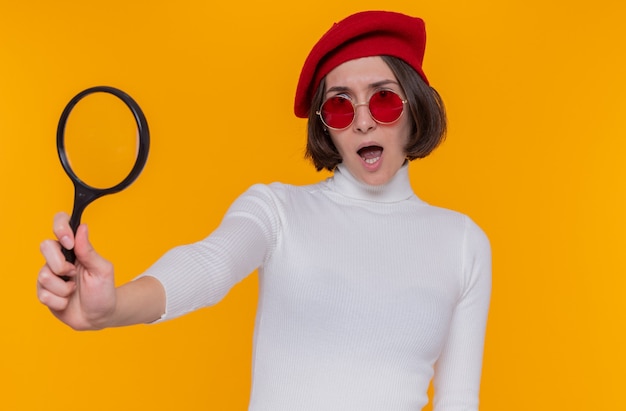 Young woman with short hair in white turtleneck wearing beret and red sunglasses holding magnifying glass looking surprised and displeased standing over orange wall