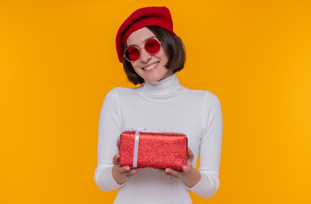 young woman with short hair in white turtleneck wearing beret and red sunglasses holding a present happy and positive