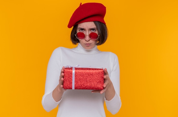 Young woman with short hair in white turtleneck wearing beret and red sunglasses holding a present looking at front with frowning face being offended standing over orange wall