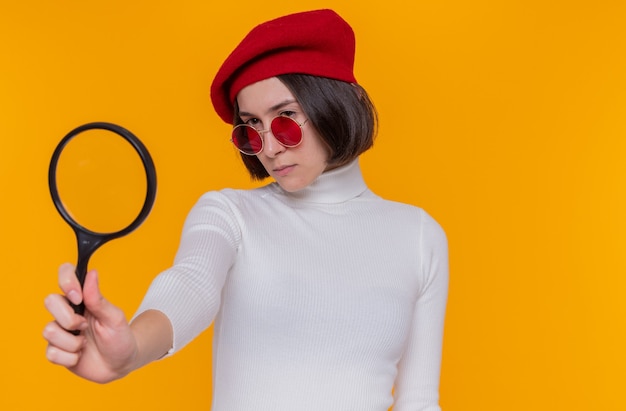 Young woman with short hair in white turtleneck wearing beret and red sunglasses looking aside through this magnifying glass with serious face standing over orange wall
