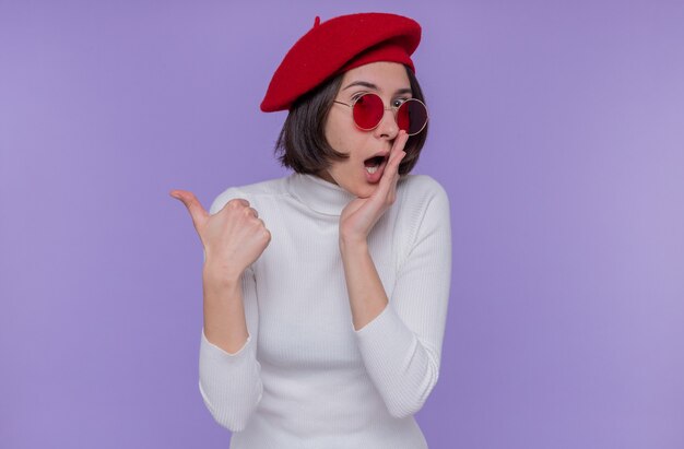 Young woman with short hair in white turtleneck wearing beret and red sunglasses looking surprised telling a secret holding hand near mouth pointing back with thumb standing over blue wall
