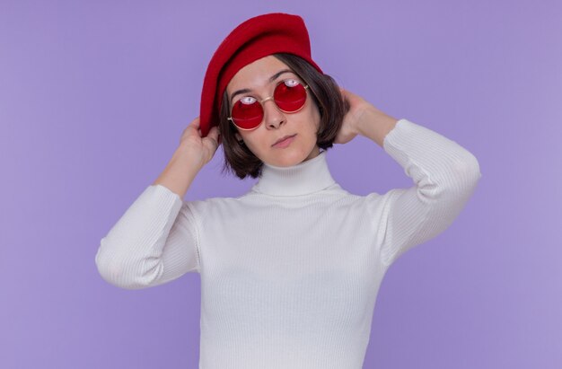 Young woman with short hair in white turtleneck wearing beret and red sunglasses looking up puzzled standing over blue wall