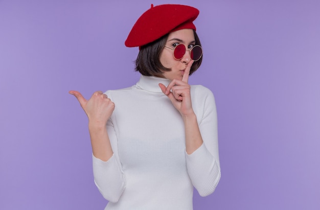 ベレー帽と赤いサングラスを身に着けている白いタートルネックの短い髪の若い女性は、青い壁の上に立っている正面を見て親指で後ろを指している唇に指で沈黙のジェスチャーをしています