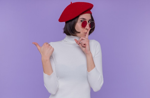 Free photo young woman with short hair in white turtleneck wearing beret and red sunglasses making silence gesture with finger on lips pointing back with thumb looking at front standing over blue wall