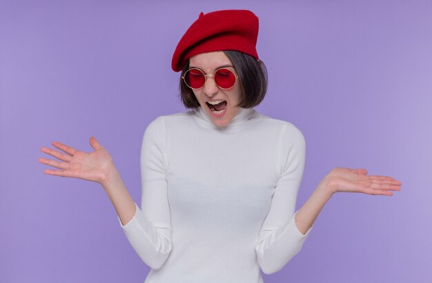 Young woman with short hair in white turtleneck wearing beret and red sunglasses screaming happy and excited raising hands standing over blue wall