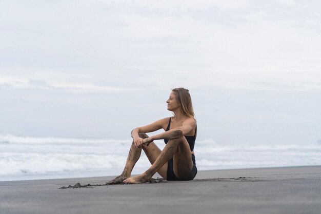 A young woman with a slender body sits on the sand by the ocean.