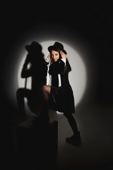 Молодая женщина со стройным телом в черной модной шляпе и коротком черном платье позирует в прожекторном черном ...