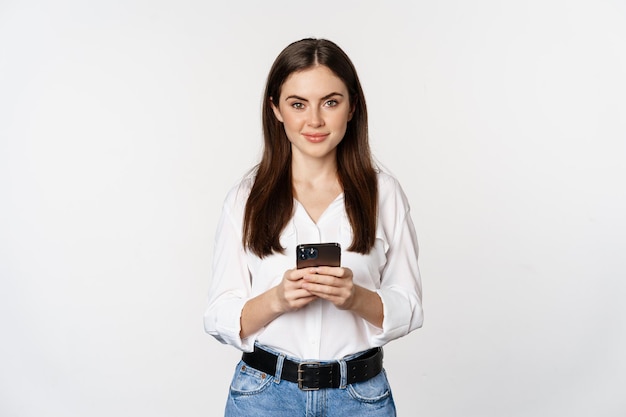 Young woman with smartphone, smiling and looking at camera, using mobile phone app, cellular technology and online shopping concept, white background
