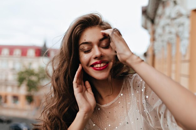 Young woman with sparkles on face smiles sincerely Attractive girl with red lips closes eyes and poses on balcony outside