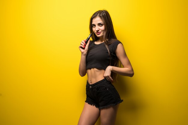 Молодая женщина с гаджетом vape курить стоя на желтой стене