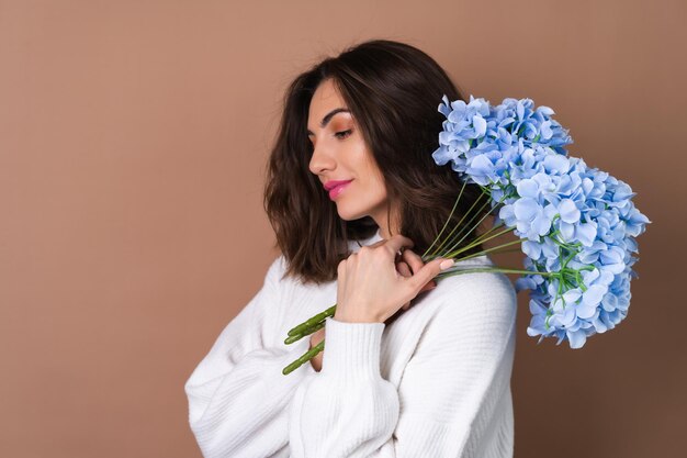 흰색 스웨터에 밝은 분홍색 립스틱 립글로스가 있는 베이지색 배경에 물결 모양의 볼륨 있는 머리를 한 젊은 여성이 파란 꽃 꽃다발을 들고 있습니다