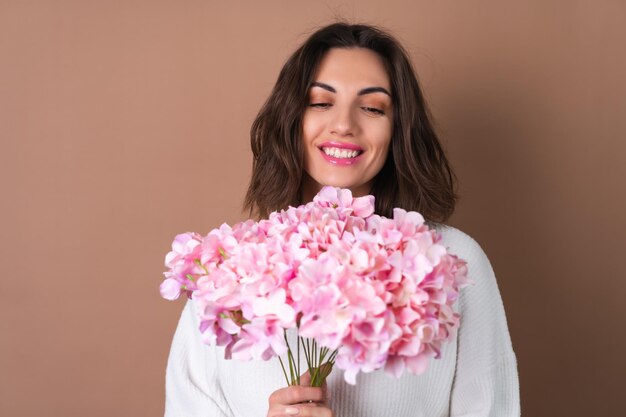 a young woman with wavy voluminous hair on a beige background with bright pink lipstick lip gloss in a white sweater holds a bouquet of pink flowers