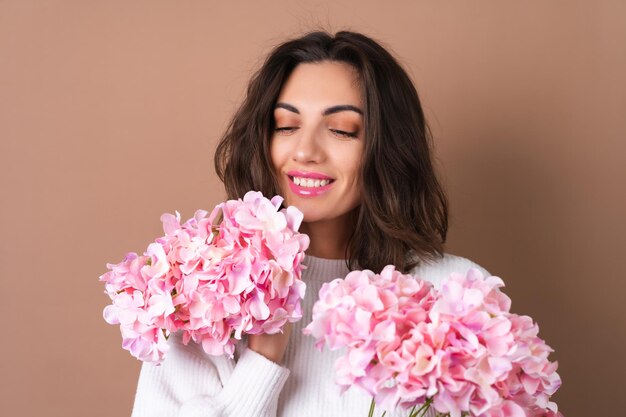 a young woman with wavy voluminous hair on a beige background with bright pink lipstick lip gloss in a white sweater holds a bouquet of pink flowers