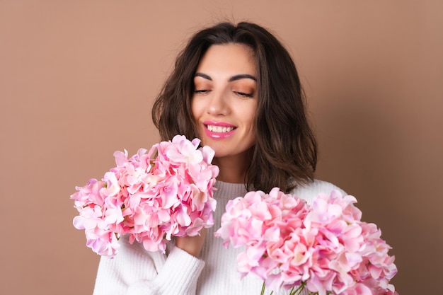 A young woman with wavy voluminous hair on a beige background with bright pink lipstick lip gloss in a white sweater holds a bouquet of pink flowers