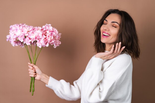 A young woman with wavy voluminous hair on a beige background with bright pink lipstick lip gloss in a white sweater holds a bouquet of pink flowers
