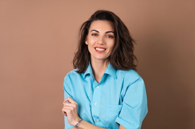 Young woman with wavy voluminous hair and natural daytime makeup wearing a blue shirt on a beige background smiles with a mischievous cheerful smile