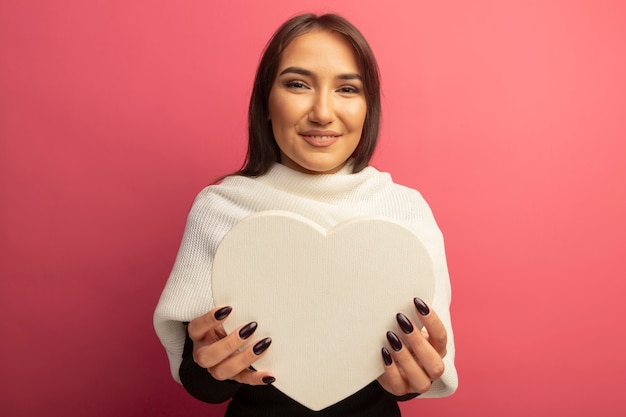 Молодая женщина с белым шарфом держит картонное сердце, искренне улыбаясь
