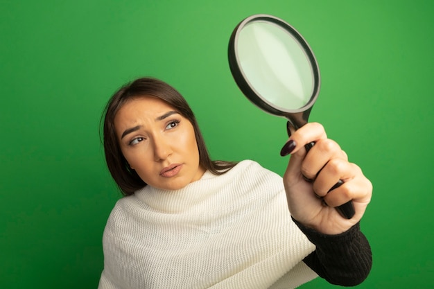 Young woman with white scarf looking at magnifying glass with serious face 