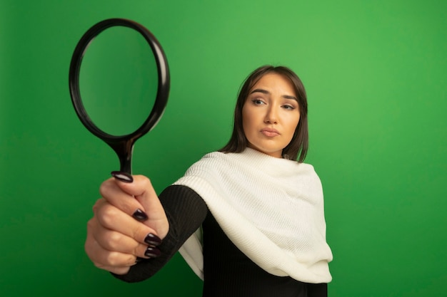 Young woman with white scarf looking at magnifying glass with serious face 