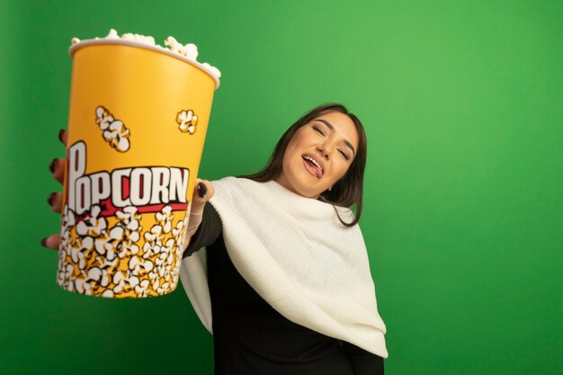 Молодая женщина с белым шарфом показывает ведро с попкорном, счастливая и веселая, высунув язык