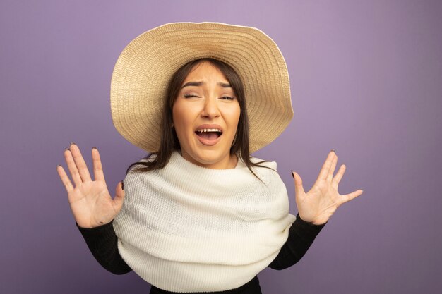 Young woman with white scarf and summer hat looking at front shouting with raised hands being scared standing over purple wall