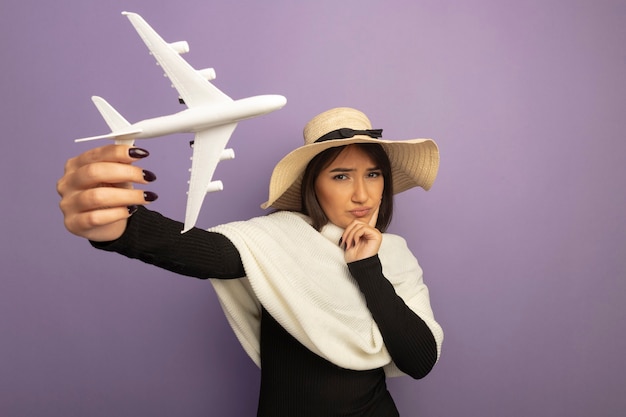 Молодая женщина с белым шарфом в летней шляпе показывает игрушечный самолет с рукой на подбородке, думая с серьезным лицом