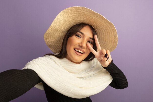 Young woman with white scarf and summer hat smiling cheerfully showing v-sign 