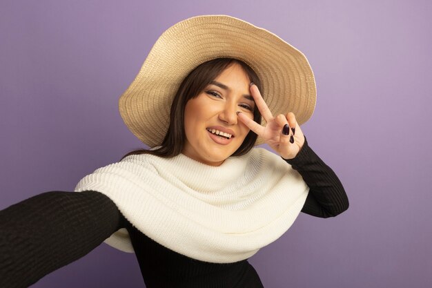 Young woman with white scarf and summer hat smiling with happy face showing v-sign 