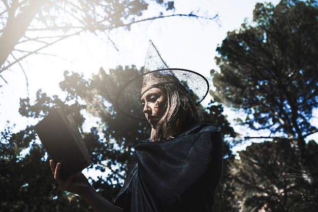 Giovane donna in cappello da mago e libro di lettura mantello nella foresta