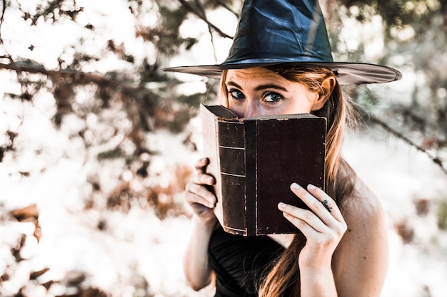 Free photo young woman in wizard hat closing mouth with book in forest