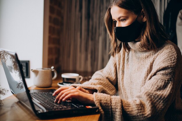 Молодая женщина, работающая на ноутбуке в кафе в маске