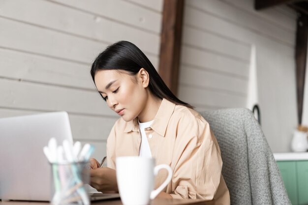 宿題を書き留めて、コンピューターでウェビナーを聞いている家の女の子でラップトップに取り組んでいる若い女性...