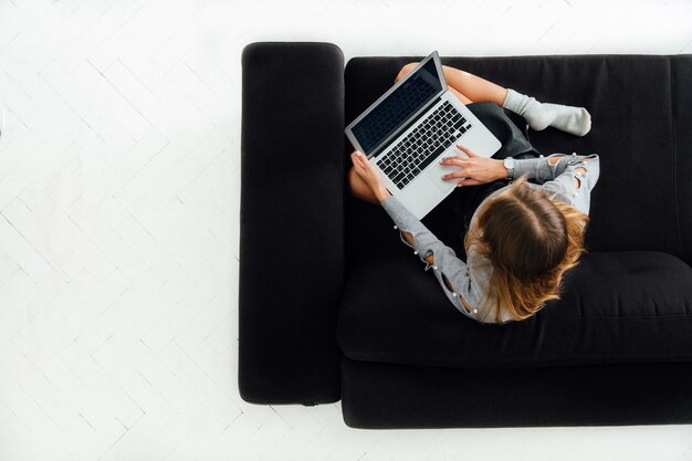 Young woman working on laptop, sitting on black cozy sofa, white floor. 
