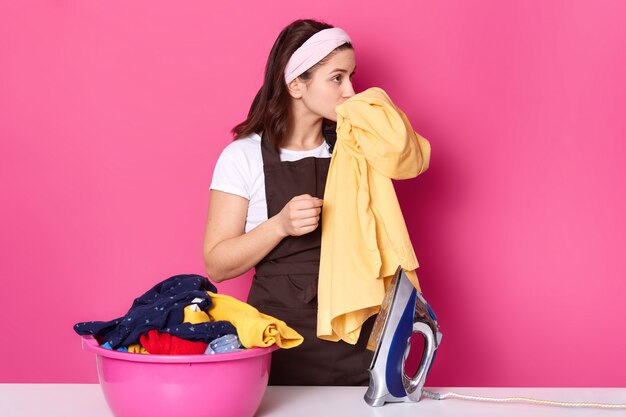 Young woman works as maid, wears t shirt, brown apron and hair band, standing near pink basin with clean linen isolated on rose in photo studio, smells fresh clothes, enjoing pleasant odor.