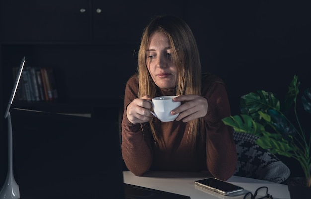 A young woman works behind a laptop at night with a lamp light