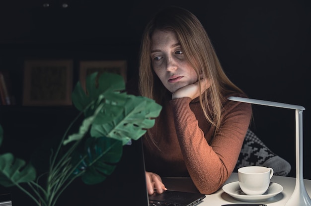 A young woman works behind a laptop at night with a lamp light