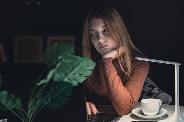 A young woman works behind a laptop at night with a lamp light