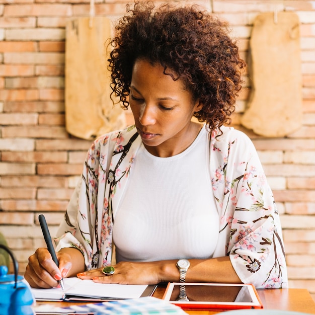 Scrittura della giovane donna in diario con la penna e la compressa digitale sulla tavola di legno