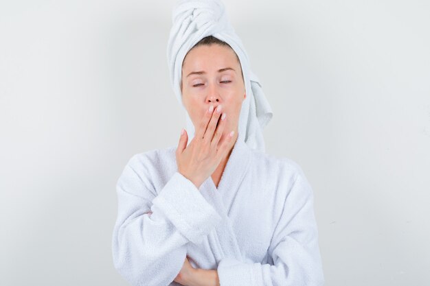 Young woman yawning in white bathrobe, towel and looking sleepy , front view.