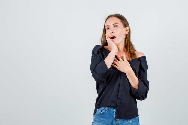 Young woman yawning with hands on chest and cheek in shirt, shorts front view.