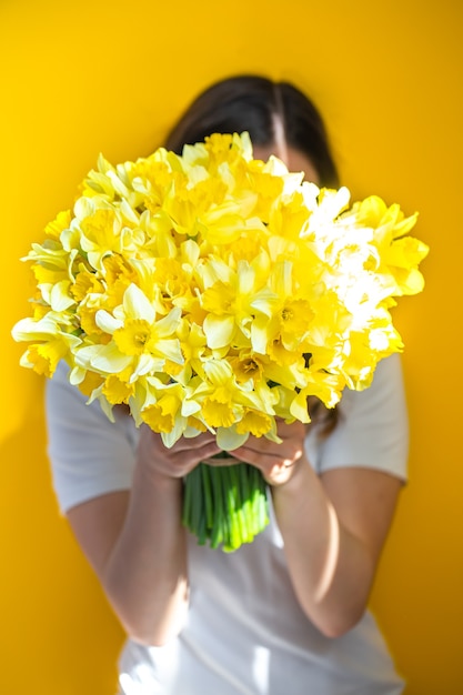 背景が黄色の若い女性は、黄色い水仙の花束で顔を覆っています。女性の日のコンセプトです。