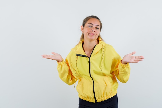 Free photo young woman in yellow bomber jacket and black pants stretching hands in questioning manner and looking puzzled