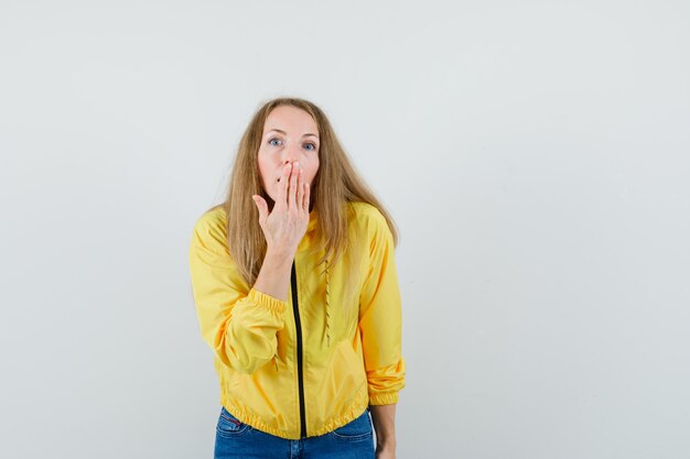 Young woman in yellow bomber jacket and blue jean covering mouth with hand and looking surprised , front view.