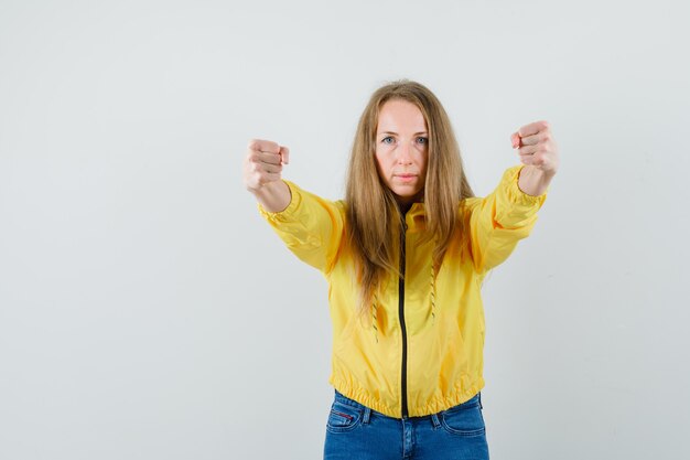 Young woman in yellow bomber jacket and blue jean holding fists clenched and looking serious
