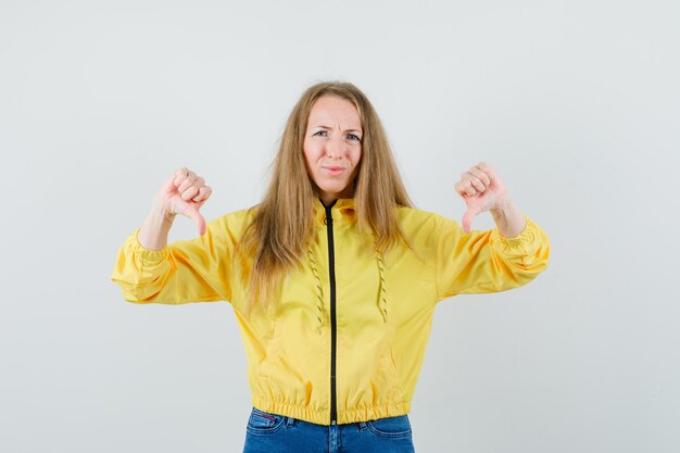 Молодая женщина в желтой куртке-бомбардировщике и синих джинсах показывает двойные пальцы вниз и недовольна, вид спереди.