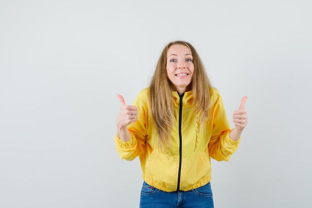 Young woman in yellow bomber jacket and blue jean showing double thumbs up and looking optimistic , front view.